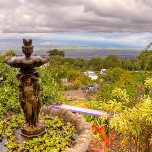 A stone fountain with a figure at its base, surrounded by lush greenery and a scenic view under a cloudy sky.