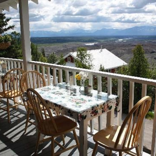 A scenic outdoor dining area with a table and chairs overlooking a valley and mountains under a cloudy sky.