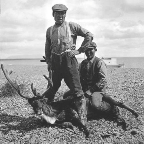 Two men pose with a reindeer on a rocky shore, with a boat visible in the background under a cloudy sky.