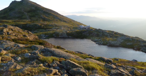 A serene mountain landscape with a small pond, rocky terrain, and a distant building under a soft sunset sky.