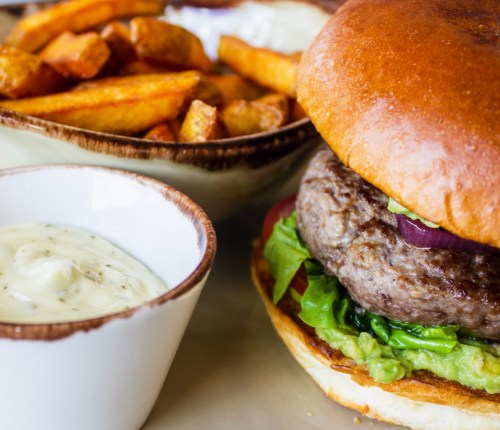 A close-up of a juicy burger with lettuce and guacamole, served with crispy fries and a small bowl of dipping sauce.