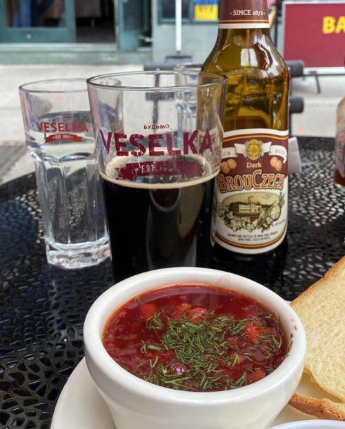 A bowl of red soup garnished with herbs, a glass of dark beer, and a bottle on a table outside a restaurant.