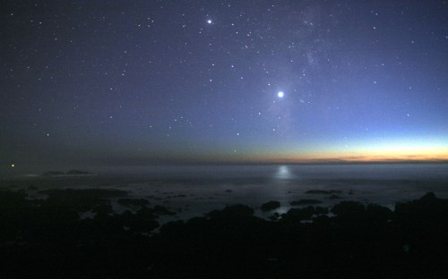 A serene night sky filled with stars over a calm ocean, with a faint horizon glow and rocky shoreline.