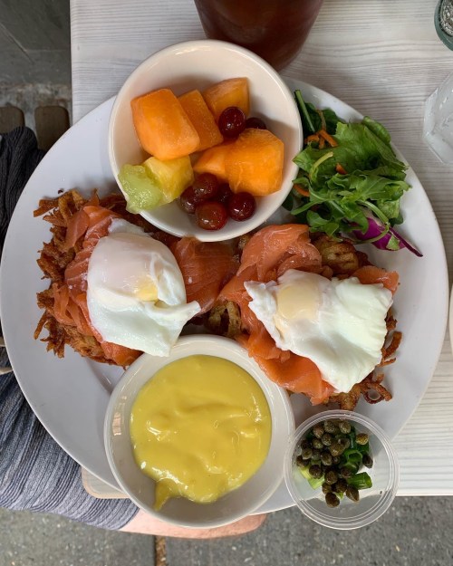 A plate with two poached eggs on smoked salmon, hash browns, fruit, salad, and a small bowl of mustard and capers.
