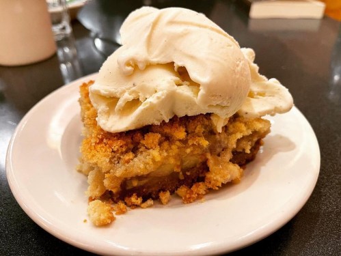 A slice of warm dessert topped with a scoop of vanilla ice cream on a white plate.