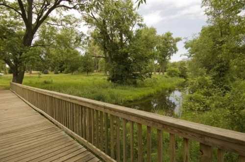 A wooden walkway leads through lush greenery beside a calm stream under a cloudy sky.