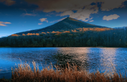 A serene lake reflects a mountain with autumn foliage under a blue sky with scattered clouds.