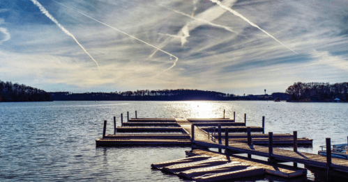 A serene lake view with a wooden dock extending into the water, under a sky streaked with clouds and contrails.