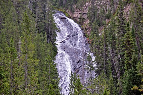 A cascading waterfall flows down rocky terrain, surrounded by lush green trees and rugged cliffs.