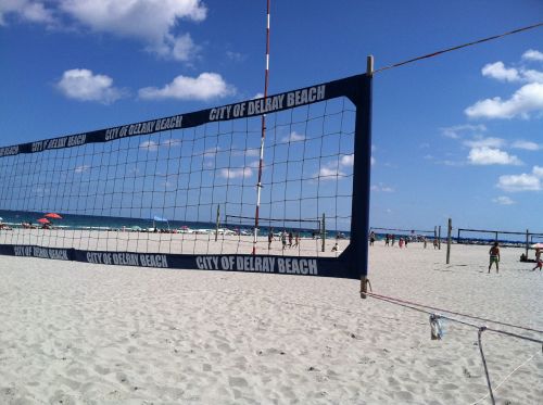 A beach volleyball court with a net, overlooking a sandy beach and ocean under a clear blue sky.