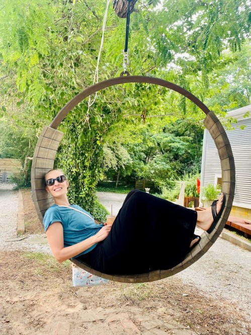 A woman relaxes on a circular swing in a lush outdoor setting, wearing sunglasses and a casual outfit.