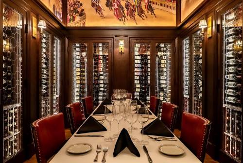 Elegant dining room with a long table set for a meal, surrounded by wine racks and a mural of horse racing.