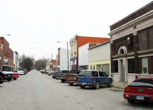A quiet street lined with parked cars and small storefronts, featuring a mix of brick and wooden buildings.