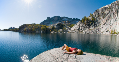 A person relaxes on a rock by a serene lake, surrounded by mountains and clear blue skies. Sunlight shines brightly.