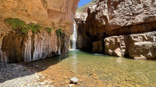 A serene canyon with a waterfall cascading into a clear pool, surrounded by rocky cliffs and lush greenery.
