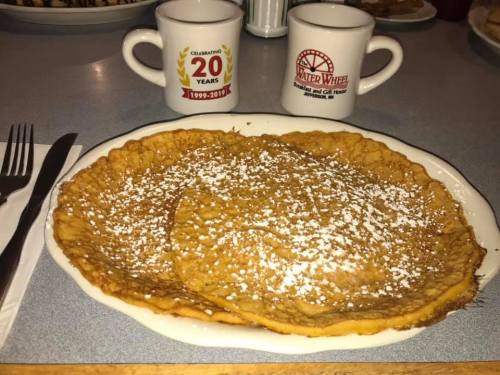 Two large, golden pancakes dusted with powdered sugar on a white plate, with two coffee mugs in the background.