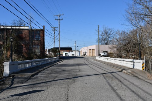 A quiet street scene featuring a bridge, power lines, and industrial buildings under a clear blue sky.