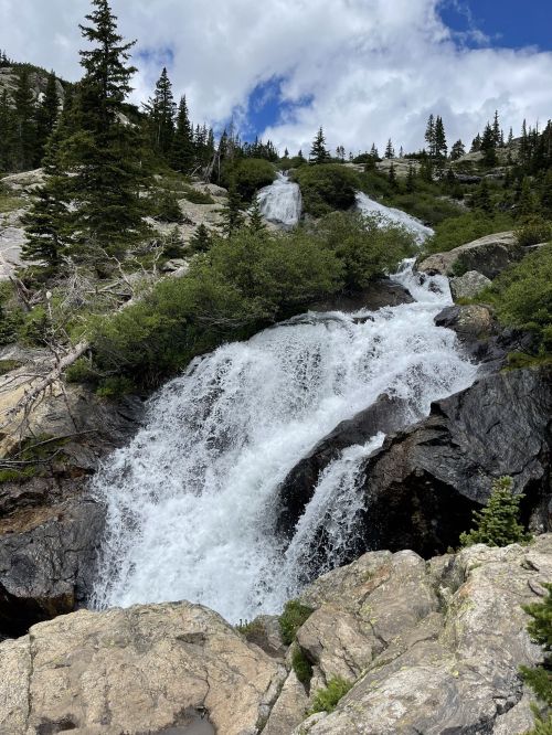 A cascading waterfall flows over rocky terrain, surrounded by lush greenery and tall pine trees under a partly cloudy sky.