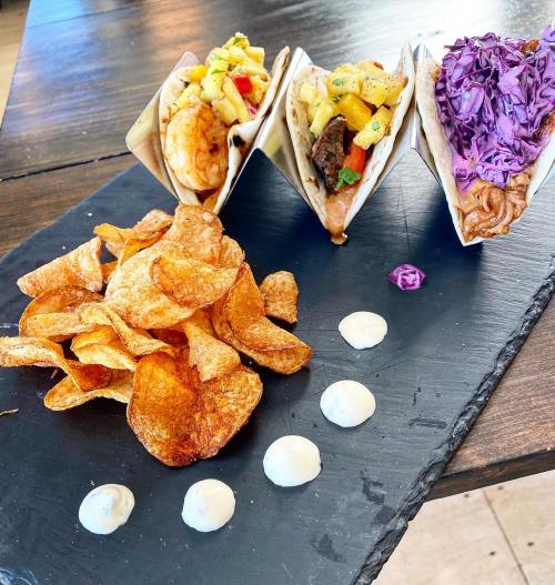 Three tacos with various fillings on a slate plate, accompanied by crispy potato chips and dollops of sauce.