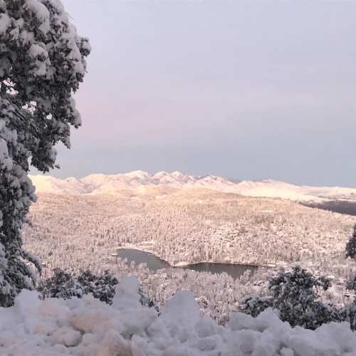 Snow-covered landscape with mountains and a lake, under a soft pink and blue sky.