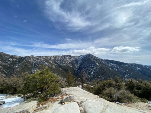 A panoramic view of rugged mountains under a partly cloudy sky, with rocky terrain and scattered trees in the foreground.