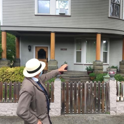A person in a suit and hat gestures towards a large, gray house with a porch and wooden columns.