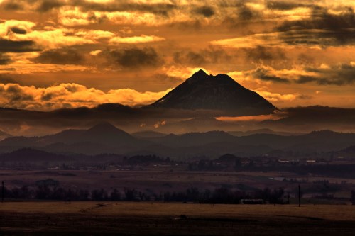 A dramatic sunset over mountains, with clouds casting shadows on a prominent peak in the distance.