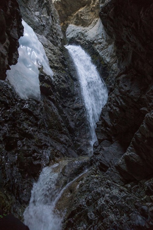 A cascading waterfall flows through a rocky crevice, surrounded by dark stone and patches of ice.