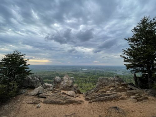 A rocky overlook with trees, showcasing a vast landscape under a cloudy sky.