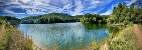 A serene lake surrounded by lush green hills and a blue sky with wispy clouds, reflecting on the water's surface.