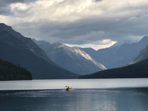 A lone kayaker paddles on a calm lake surrounded by mountains under a cloudy sky.