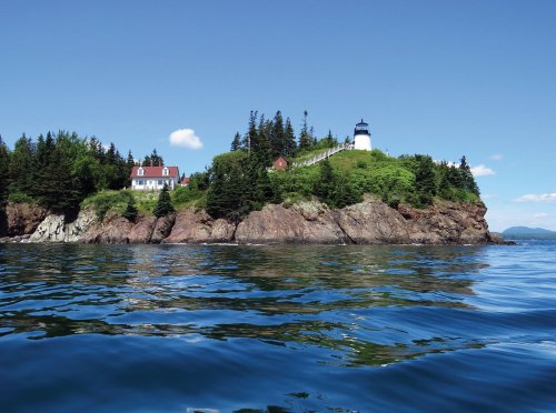 A scenic view of a lighthouse on a rocky shore, surrounded by lush greenery and calm blue waters under a clear sky.