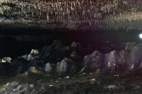 A dark cave interior with rocky formations and stalactites hanging from the ceiling, illuminated by faint lights.