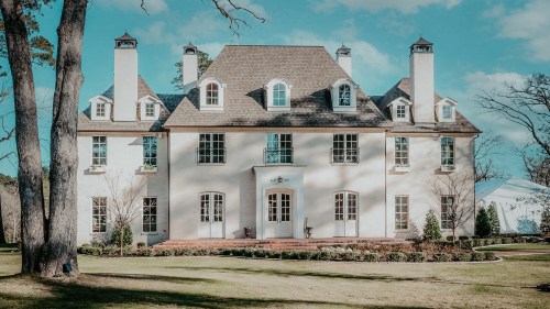 A large, elegant two-story house with a steep roof, multiple windows, and a manicured lawn under a blue sky.