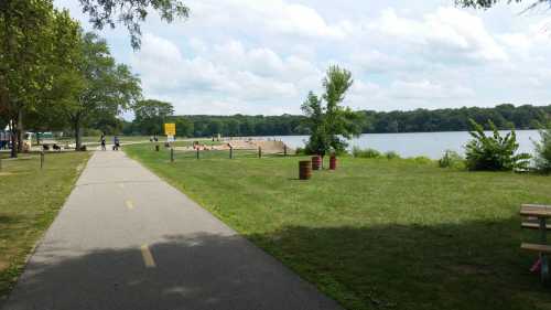 A paved path leads to a beach area by a lake, surrounded by trees and grassy fields under a cloudy sky.