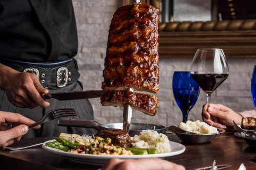 A server carves a large rack of ribs at a dining table with sides of mashed potatoes and asparagus, accompanied by wine.