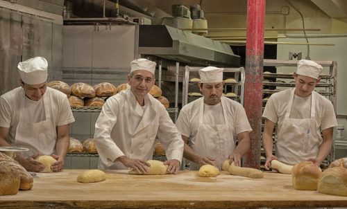 Four bakers in white uniforms work at a wooden table, shaping dough in a bakery filled with loaves of bread.