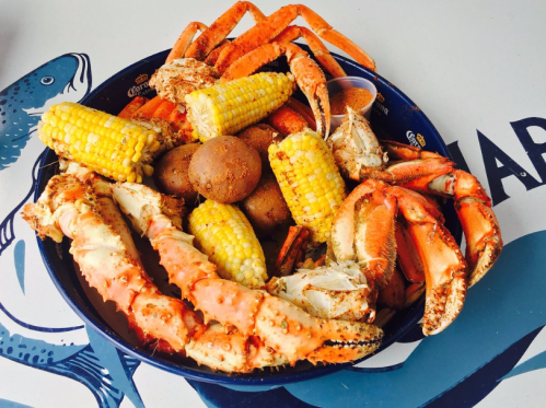 A colorful seafood platter featuring crab legs, corn on the cob, and round bread rolls on a blue table.
