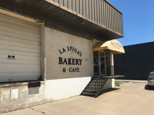 Exterior of La Spiga's Bakery & Café, featuring a yellow awning and a loading dock area. Clear blue sky above.