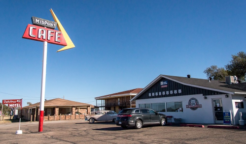 Sign for Midpoint Cafe with a retro design, alongside a building and parked cars under a clear blue sky.
