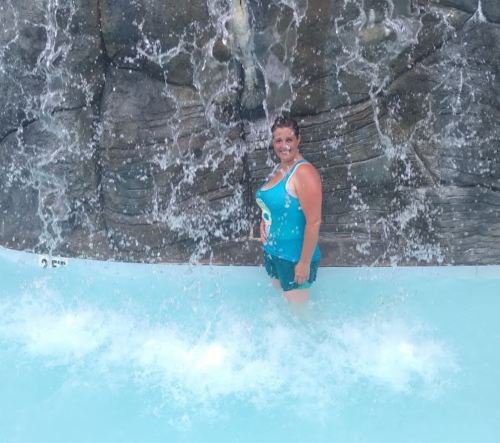 A woman stands in a pool, smiling, with a waterfall cascading behind her. Water splashes around her.