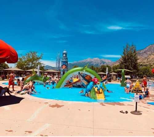 A vibrant water park scene with families enjoying a pool, slides, and playful dragon sculptures under a clear blue sky.
