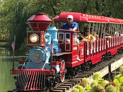 A colorful steam train with passengers rides along a track beside a pond, surrounded by greenery.