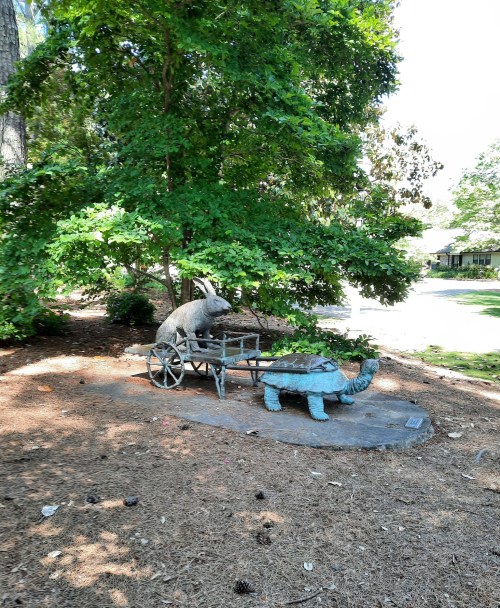 A bronze sculpture of a rabbit pulling a cart with a turtle, surrounded by greenery and pine needles.