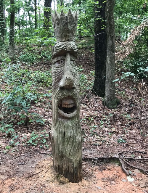 A carved wooden statue of a face with a crown, surrounded by trees in a forested area.