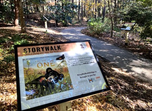 A path in a wooded area with a sign for a StoryWalk featuring the book "One" and nature-themed illustrations.