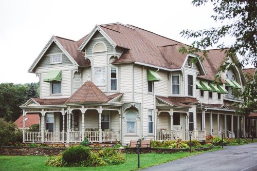 A charming Victorian-style house with a wraparound porch and green awnings, surrounded by lush landscaping.