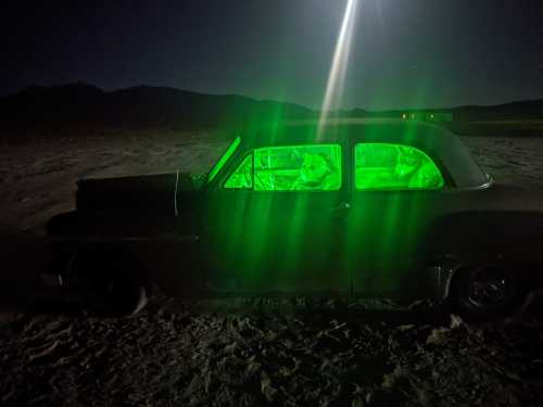 A vintage car illuminated with green light at night, parked in a desert landscape with mountains in the background.