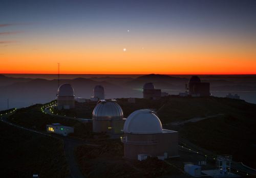 A scenic view of observatories at sunset, with a vibrant orange sky and distant mountains.