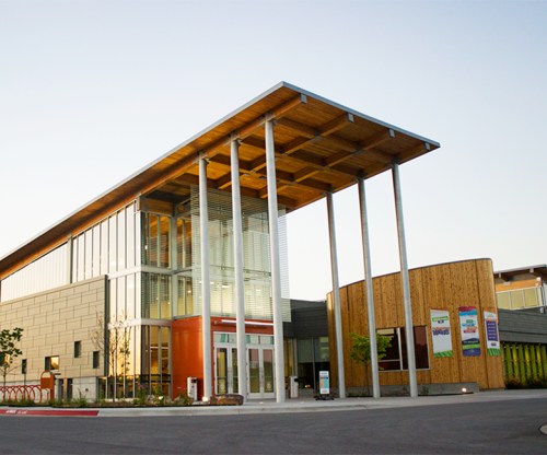 Modern building with a wooden roof and large glass entrance, featuring tall columns and a landscaped area.